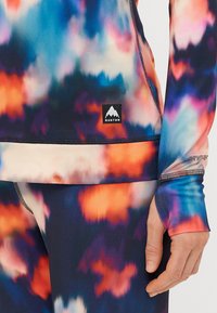 Blusa de mangas compridas colorida e calças combinando feitas de um tecido elástico com um padrão degradê em azuis, laranjas e roxos. Apresenta logotipo e buracos para o polegar.