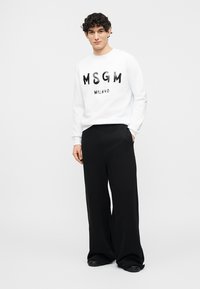 Άνδρας που φοράει λευκό φούτερ MSGM Milano και φαρδιά μαύρη παντελόνα, στέκεται με το ένα χέρι στην τσέπη μπροστά σε απλό φόντο.