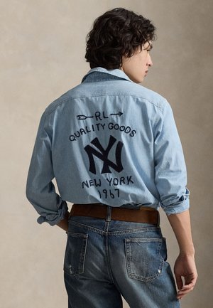 Lys blå denimskjorte med brodert logo på baksiden, med teksten «RL QUALITY GOODS» og «NEW YORK 1967». Brukt sammen med blå jeans.