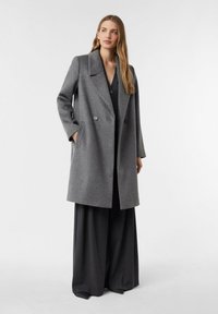 Szary dwurzędowy płaszcz z mieszanki wełny z wyciętym kołnierzem, bocznymi kieszeniami i dwoma dużymi guzikami. Modelka ma na sobie ciemny strój pod spodem.