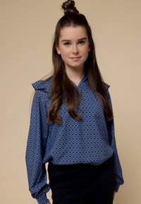 Blauwe blouse met patroon en kraag, met diamantvormen. Lange mouwen met elastische manchetten. Draag over zwarte onderkleding.