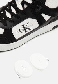 Sneaker in pelle bianca e nera con pannelli texturizzati, suola bianca, lacci neri, logo Calvin Klein Jeans e due lacci bianchi arrotolati.
