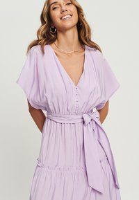 Lavanda lepršava obleka z V-izrezom, kratkimi rokavi, podrobnostmi gumbov ter pasom. Ima večplastno krilo in mehak, lahek material.