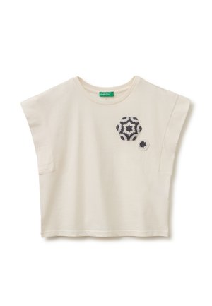 Chemise crème à manches courtes avec manches larges et deux patchs étoilés hexagonaux noir et blanc sur le côté gauche de la poitrine.