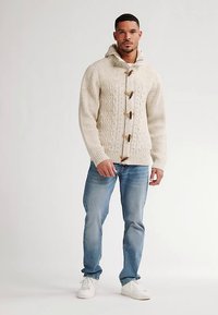 Krémový svetr s pleteným vzorem a kapucí, s dřevěnými knoflíky, kombinovaný se světle modrými džíny a bílými teniskami.