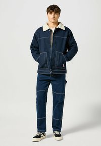Granatowa kurtka dżinsowa z kołnierzem Sherpa, z przodu na zamek błyskawiczny, dwoma kieszeniami, kontrastowymi szwami. W zestawie z pasującymi spodniami i obuwiem sportowym.