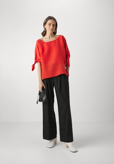 Red, loose-fit top with short sleeves and tie detail, paired with black wide-leg trousers and white heeled ankle boots. Small black handbag.