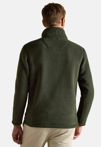 Uomo che indossa una giacca in pile verde scuro con logo in rilievo sul colletto, di spalle, abbinata a pantaloni beige su sfondo bianco.