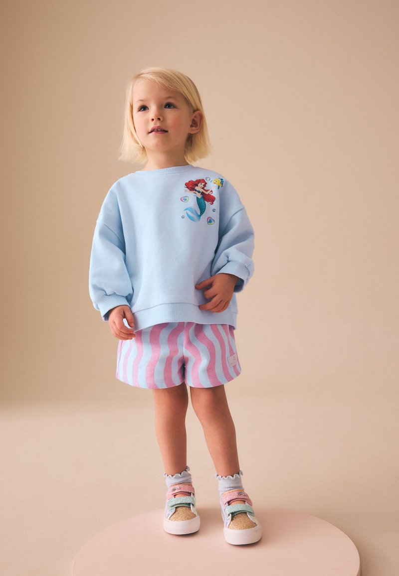Niño pequeño con pelo rubio que lleva una sudadera azul claro con estampado de sirena, pantalones cortos a rayas rosas y blancas y zapatillas de colores.