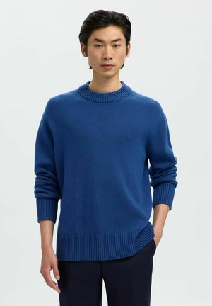 Jeune homme aux cheveux noirs portant un pull bleu à col rond avec des poignets et un ourlet côtelés, se tenant avec une main dans la poche sur fond blanc.