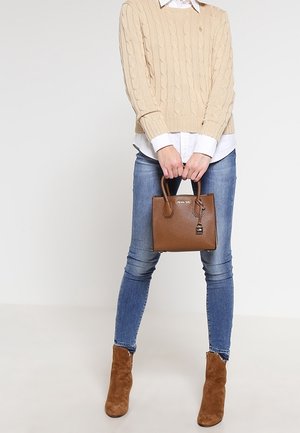 Osoba w beżowym swetrze z warkoczowym splotem, białej koszuli, niebieskich obcisłych dżinsach i brązowych botkach trzymająca brązową skórzaną torebkę.