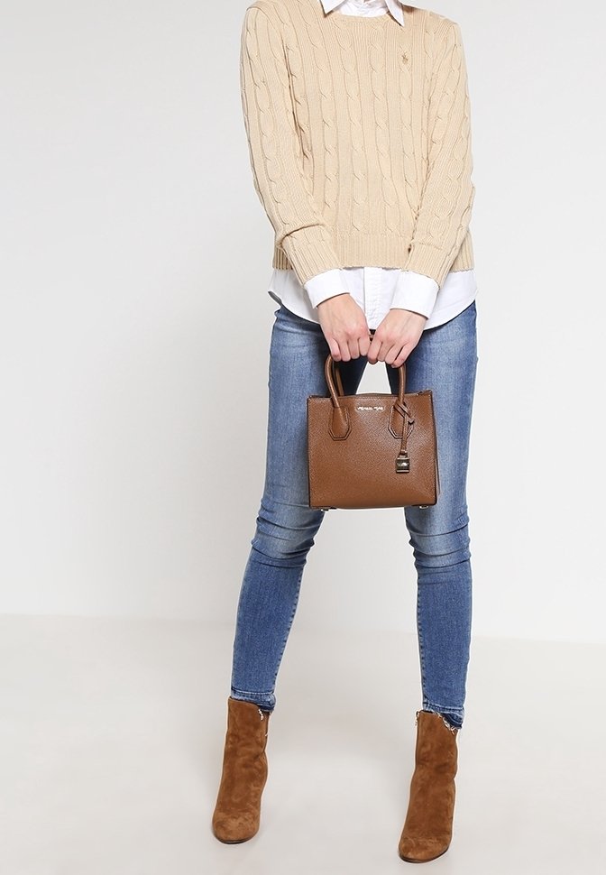 Personne en pull beige à torsades, chemise blanche, jean skinny bleu et bottines marron tenant un sac à main en cuir marron.