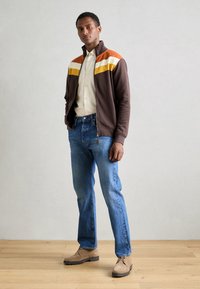 Braune Zip-Jacke mit orangefarbenen, gelben und weißen Streifen; cremefarbenes Hemd; blaue Jeans; und beiges Wildleder-Schuhwerk.