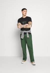 Maglietta nera con logo grafico, felpa grigia legata in vita, pantaloni verdi e sneakers beige. Tessuto morbido con una vestibilità comoda.
