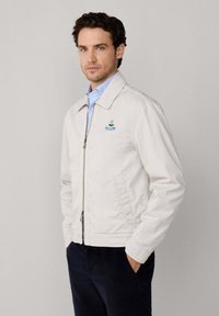 Hombre con ojos azules que lleva una chaqueta cremoso claro con cremallera sobre una camisa azul clara, con las manos en los bolsillos de unos pantalones oscuros, frente a un fondo liso.