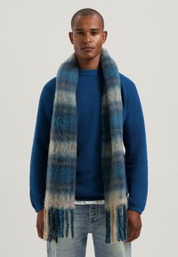 Man in een blauwe gebreide trui en een lange, fluffy blauw- en beige gestreepte sjaal, gecombineerd met lichtblauwe jeans.