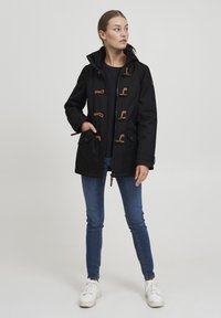 Svart regnjakke med hette, med knappefester og lommer. Kombinert med blå jeans og hvite sneakers. Glatt tekstur, slitesterkt stoff.