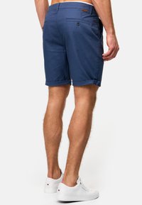 Shorts bleu marine en tissu tissé, avec ourlets retournés, une poche arrière avec fermeture par bouton, et une petite étiquette logo marron.