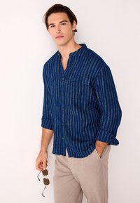 Marineblauw overhemd met knopen en witte gestreepte patronen, voorzien van een borstzak en omgeslagen mouwen. Model houdt een zonnebril vast en draagt beige broek.