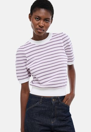 Femme aux cheveux courts portant un haut en maille à manches courtes rayé blanc et lavande et un jean bleu foncé, main dans la poche, sur un fond uni.