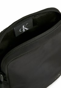 Borsa in tessuto nero con apertura con cerniera, caratterizzata da una superficie liscia e un'etichetta con logo visibile all'interno. Forma rettangolare semplice.