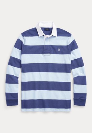 Jeune homme portant une casquette blanche, une veste en jean bleue, un polo rayé, un short rouge et des baskets bleues, debout les mains dans les poches de la veste.