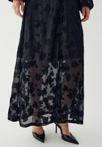 Długa czarna sukienka z przezroczystą kwiatową narzutą, ozdobiona czarnymi koralikowymi wzorami, z guzikami na przodzie i noszona z czarnymi szpilkami.