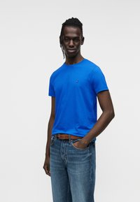 Μπλε βαμβακερό μπλουζάκι με στρογγυλή λαιμόκοψη και κοντά μανίκια, με μικρό λογότυπο στο στήθος, συνδυασμένο με ανοιχτόχρωμο τζιν παντελόνι.