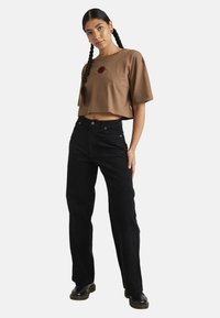 T-shirt cropped marron avec broderie rouge et jean taille haute noir. Modèle portant des bottes compensées noires, cheveux tressés.
