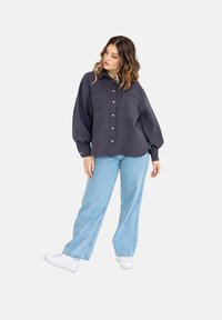 Ciemnoniebieska, oversize'owa koszula z zapięciem na guziki, dwoma kieszeniami na piersiach i długimi bufkami, w połączeniu z jasnoniebieskimi jeansami typu wide-leg oraz białymi trampkami.