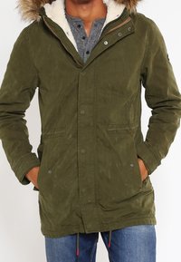 Olivgrön parkajacka med en huva foder i konstpäls, knäppt framtill, två sidofickor och dragsko i nederkant. Texturerad tyg med en avslappnad passform.