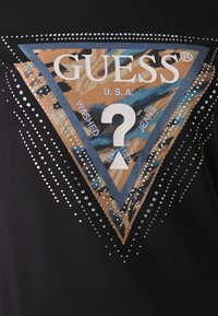 Svart t-shirt med en färgglad triangulär grafisk design med "GUESS"-logotyp, omgiven av silverfärgade rhinestones och en detalj i form av ett frågetecken.