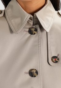 Beige-takki kolari, jossa on kolme kilpikonnankuoren nappia ja punaisia langan yksityiskohtia, jota kantaa tummatukka.