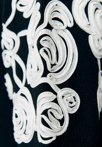 Tissu noir avec des motifs floraux brodés en relief blancs présentant des pétales et des feuilles texturés et superposés en détail rapproché.