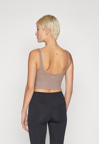 Top de desporto em tecido canelado taupe com alças largas e costas baixas; combinado com leggings pretas de cintura alta. Detalhe de logótipo discreto nas costas.