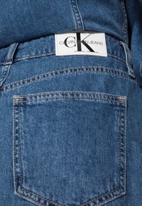 Blå denimjeans med en bakficka och en vit etikett med "Calvin Klein Jeans" och bokstäverna "CK". Strukturerad tyg med kontrasterande sömmar.