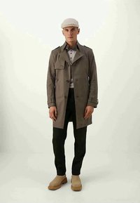 Νεαρός άνδρας με μπεζ καπέλο, γκρι παλτό, λευκό πόλο, μαύρο παντελόνι και μπεζ μπότες, που στέκεται μπροστά σε απλή ανοιχτόχρωμη φόντο.