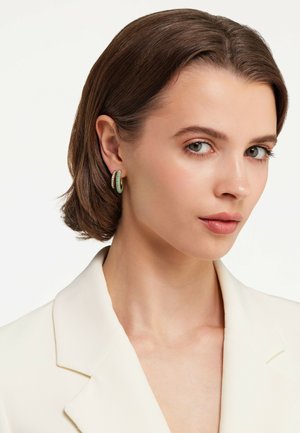 Guldörhängen i form av ringar med en grön detalj och klara stenar längs ena sidan, placerade mot en neutral bakgrund med en modell som bär en krämfärgad kavaj.