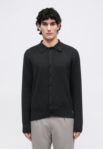 Giovane uomo con capelli neri ricci, che indossa un cardigan a costine grigio scuro con bottoni e pantaloni grigi chiari, in piedi davanti a uno sfondo neutro.