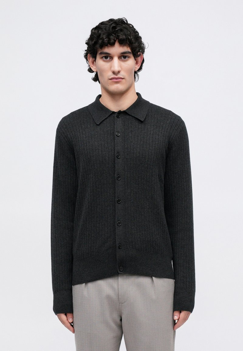 Young man with curly black hair wearing a dark gray ribbed buttoned cardigan and light gray pants, standing against a plain background.