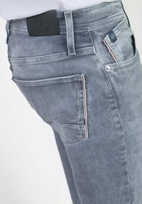 Jean en denim gris avec une texture légèrement délavée, présentant une poche arrière avec un accent de couture rouge et blanche et une étiquette de taille noire.