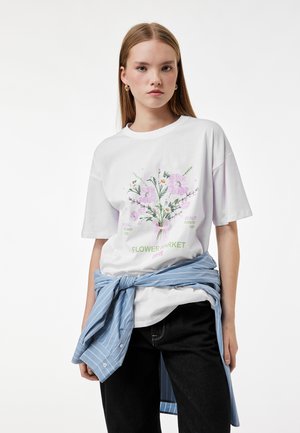 Młoda kobieta w białej koszulce z różowo-zielonym wzorem kwiatowym i napisem "Flower Market", z niebieską koszulą w paski zawiązaną wokół talii.