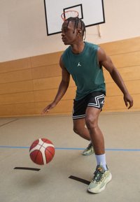Zöld ujjatlan mez logóval, fekete rövidnadrág fehér díszítéssel, és világoszöld sportcipő; kosárlabdát dribblizik egy edzőteremben.