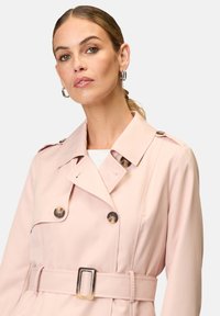 Light pink trench coat with a double-breasted design, large buttons, shoulder epaulets, and a matching waist belt. Smooth texture.