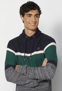 Dzianinowa bluza z kapturem w granatowe, zielone i białe paski. Posiada kołnierz z wysokim stójką, sznurek do regulacji oraz teksturowane, wzorzyste rękawy.