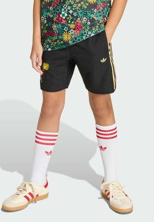 Persona con pantaloncini neri con strisce gialle e dettagli floreali, calzini bianchi con strisce rosse e scarpe da ginnastica beige con accenti rossi.