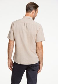 Beżowa lniana koszula z krótkim rękawem i kołnierzykiem, z plisą z tyłu i zaokrąglonym dołem. Noszona z granatowymi spodniami.