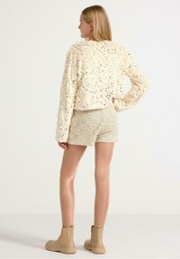 Hæklet cremet trøje med et struktureret design, parret med beige tweed shorts og lysebrune ankelstøvler. Vist fra ryggen.