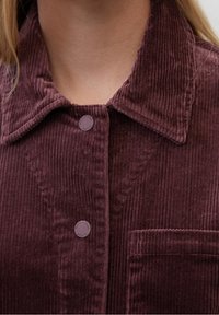Vaatteena on syvän burgundinvärinen corduroy-paita, jossa on pystyribbitys, kaulus, kaksi nappia ja rintatasku. Miellyttävä, sileä pinta, jossa on hienovarainen kiilto.