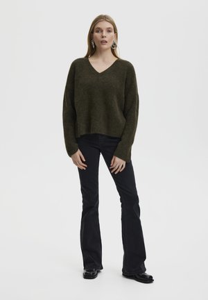 Olijfgroen v-hals sweater van zacht, textuurstof, gecombineerd met zwarte flared jeans en zwarte laarzen. Model staat tegen een effen achtergrond.